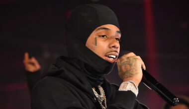 Pooh Shiesty wearing a black hood and holding a microphone performs on stage. They have a teardrop tattoo and visible jewelry.