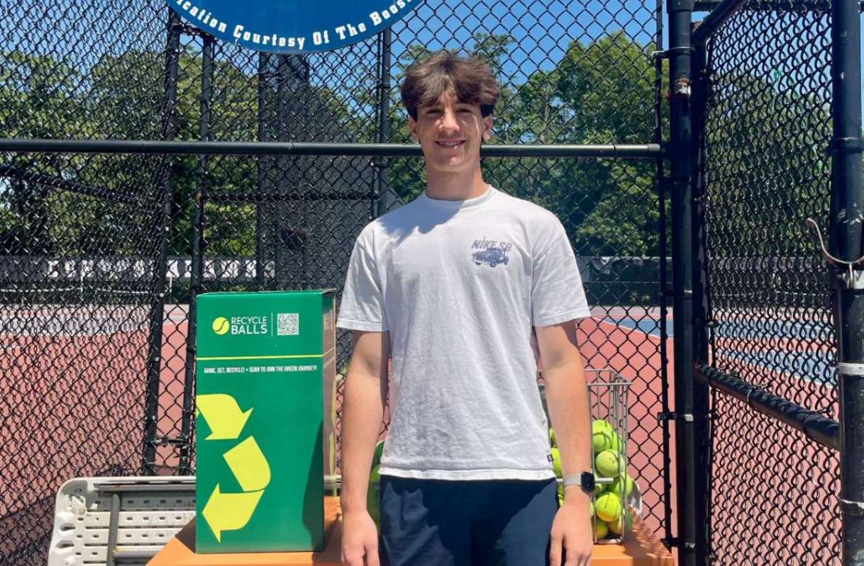 A young man stands beside a green recycling bin for used tennis balls at a tennis court.