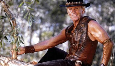 Paul Hogan as Mick Dundee in a leather vest and akubra sitting in a tree