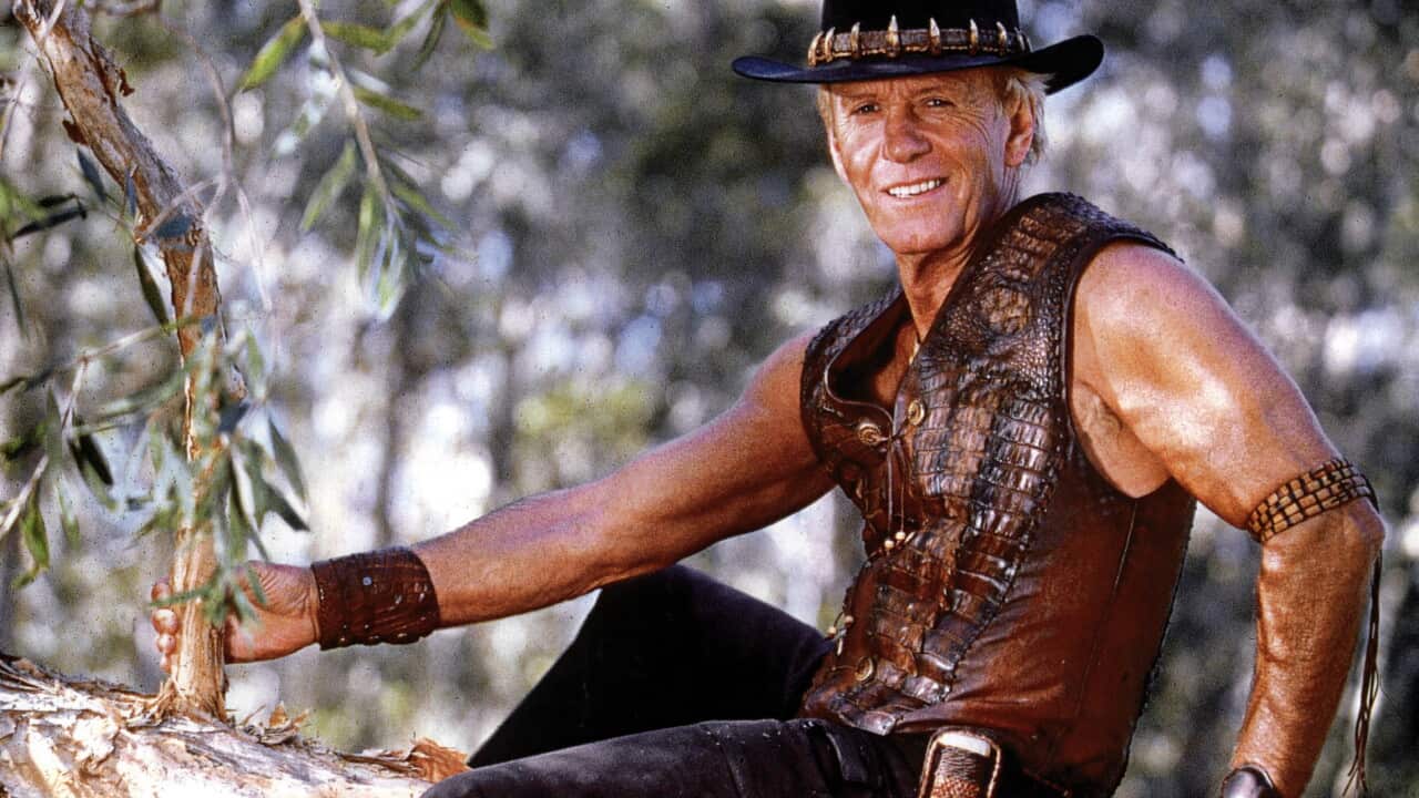 Paul Hogan as Mick Dundee in a leather vest and akubra sitting in a tree