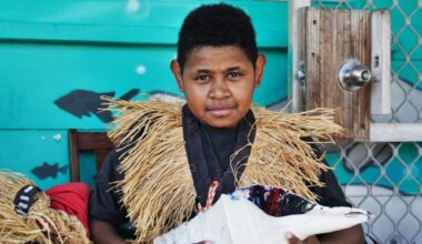 A young boy wearing a dark t-shirt, a light-brown coloured grass garment around his neck and holding a large white conch shell