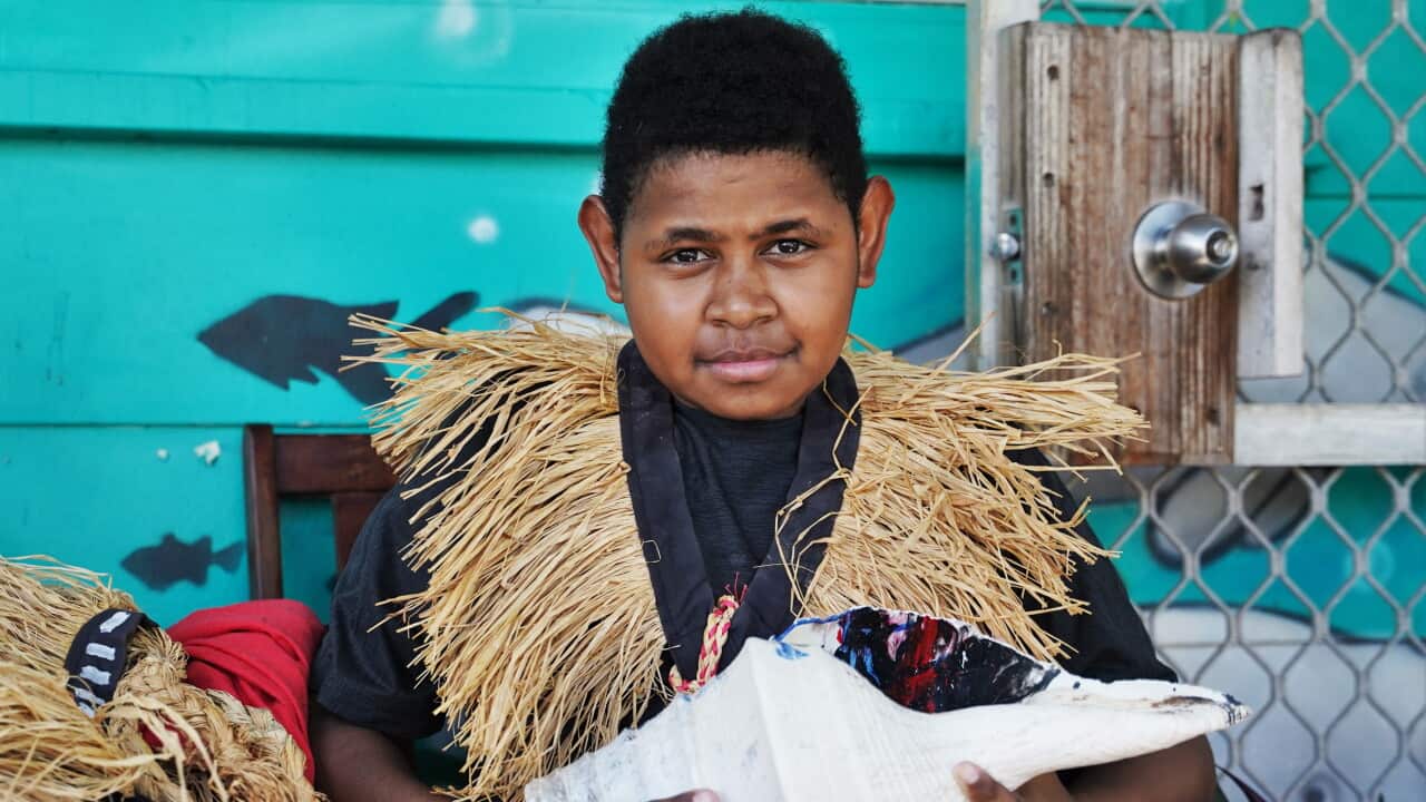 A young boy wearing a dark t-shirt, a light-brown coloured grass garment around his neck and holding a large white conch shell