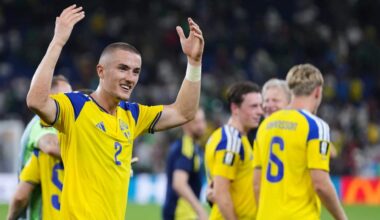 A smiling Swedish soccer player wearing a yellow and blue jersey with the number 2 raises his hands to celebrate or wave on the field alongside teammates.