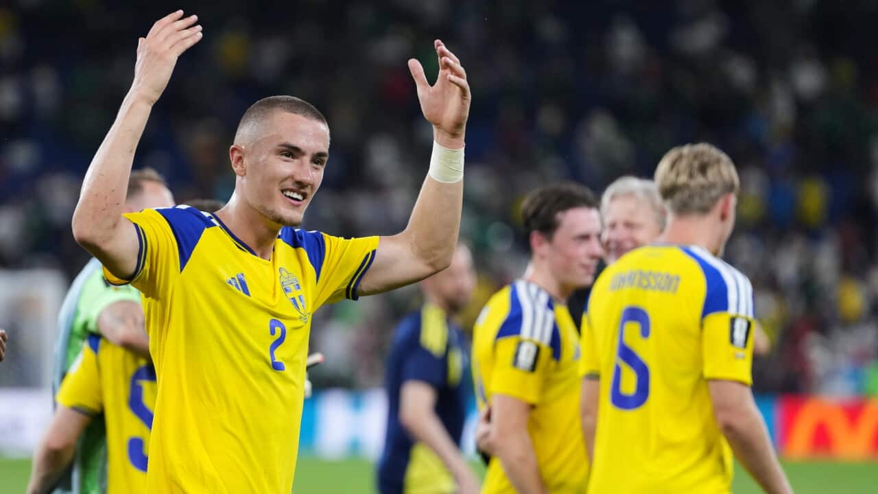 A smiling Swedish soccer player wearing a yellow and blue jersey with the number 2 raises his hands to celebrate or wave on the field alongside teammates.
