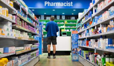 A man in a blue shirt and black shorts stands at a pharmacist's counter.
