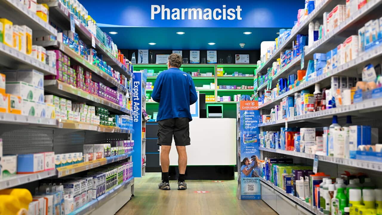 A man in a blue shirt and black shorts stands at a pharmacist's counter.