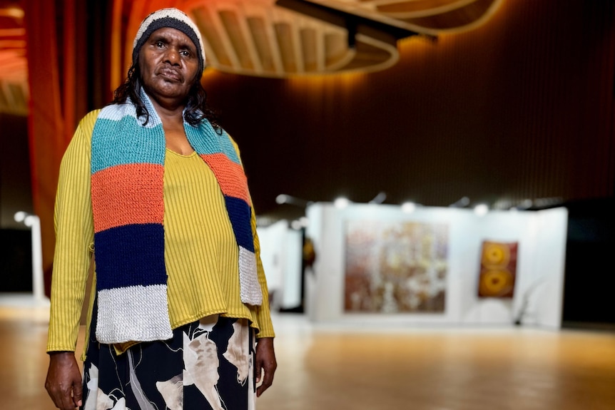 A person wearing a yellow jumper, colourful scarf and knitted hat stands in a large room with paintings in the background.