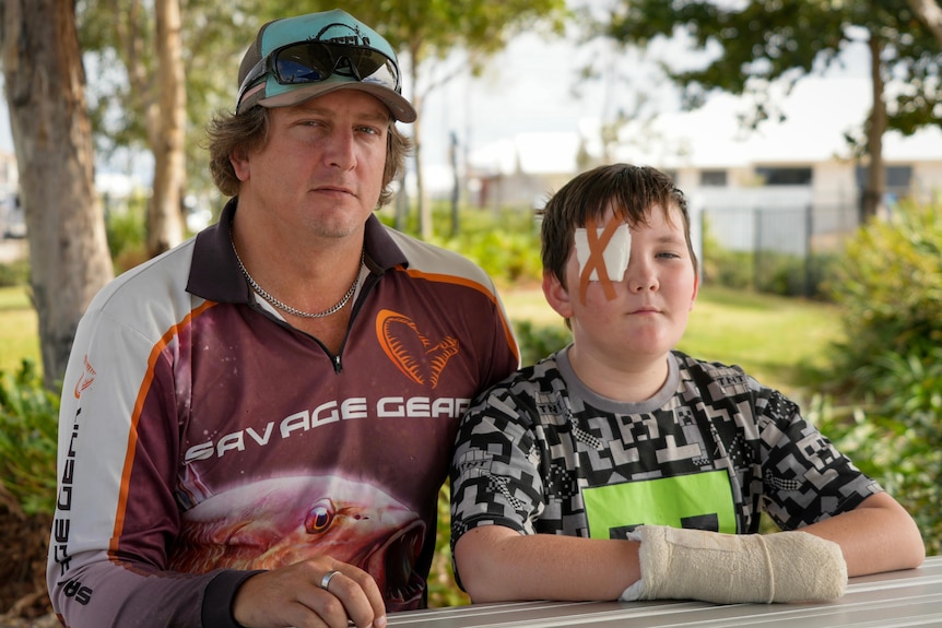 A man in a fishing shirt and cap sits at a park table with his son, who has an eye patch and an arm cast