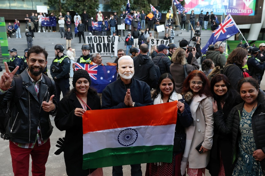 Seven people smile for the camera holding up an Indian flag while people with 'Modi go home' signs stand behind