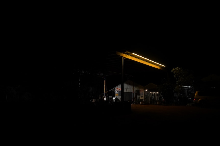 A dark image of the exterior of an outback tin shed hotel, with a yellow row of lights on the shelter roof offering dim light.