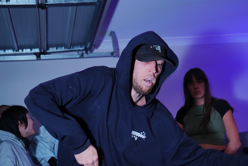 A man with stubble and a wearing a hoodie swaying his body during a dance session in a suburban garage.