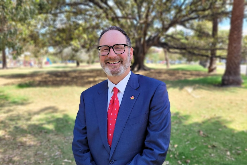 A man in a blue suit and red tie stands in a park