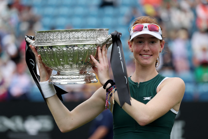 A smiling red-headed female tennis player holds big silver trophy in front of crowd