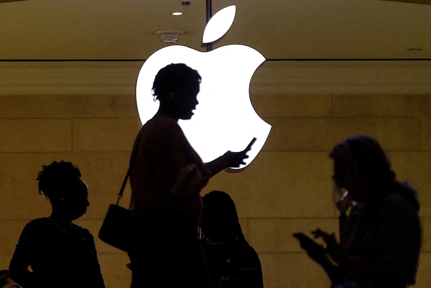 A womanan uses her phone as she passes a large and illuminated Apple logo