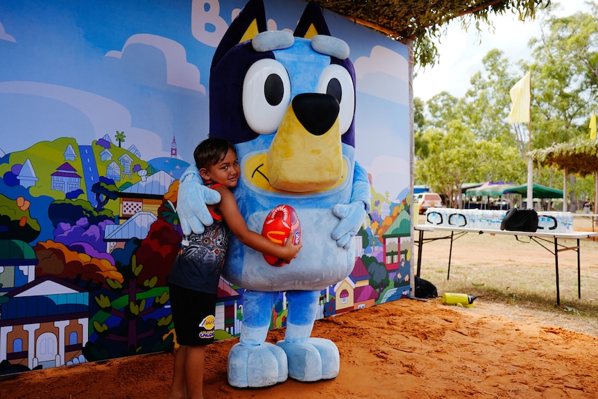 A smiling, dark-haired boy cuddles a person in a Bluey suit in front of a backdrop in the bush.