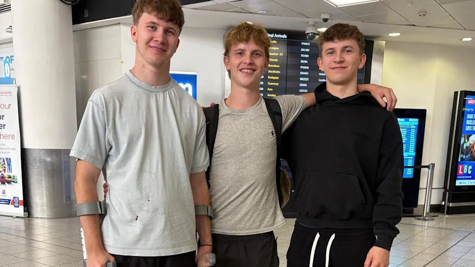 Three young men pose together in an airport lounge. The man on the left is using two crutches to support himself.