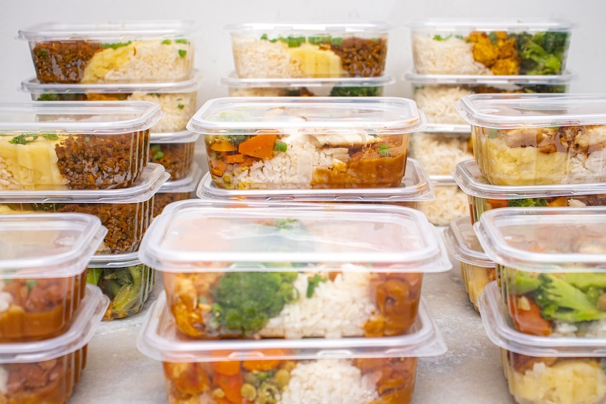 Meals of meat and vegetables in sealed plastic containers, stacked in piles on a bench.