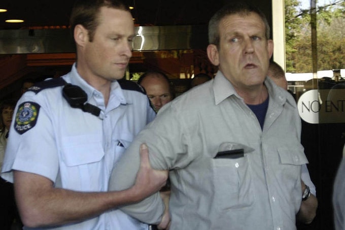 A man in a pale shirt is held by a serious-looking police officer.
