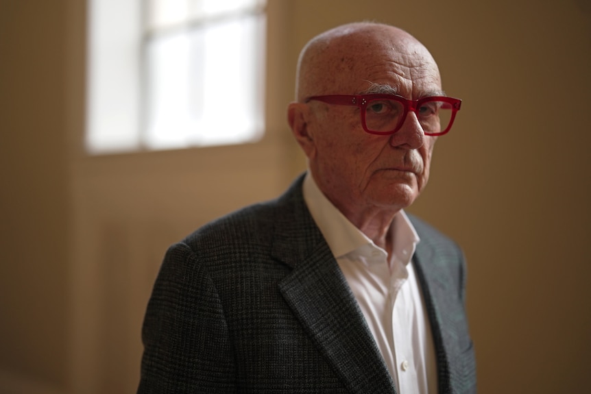A man wearing glasses stands indoors looking to the camera with a serious expression.