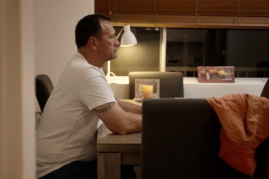 Man in a white tshirt sits at a kitchen table looking ahead.