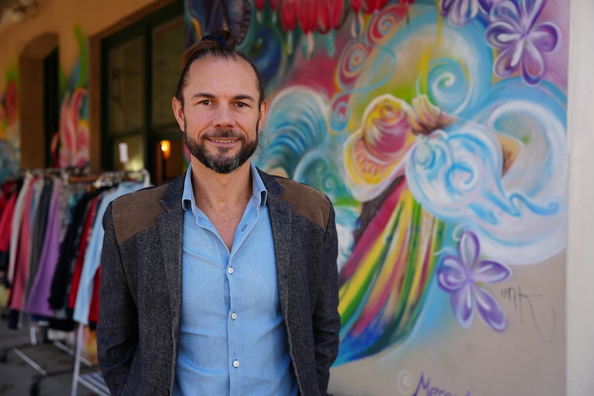 Man in a blue business shirt and grey jacket standing near a colourful wall mural.