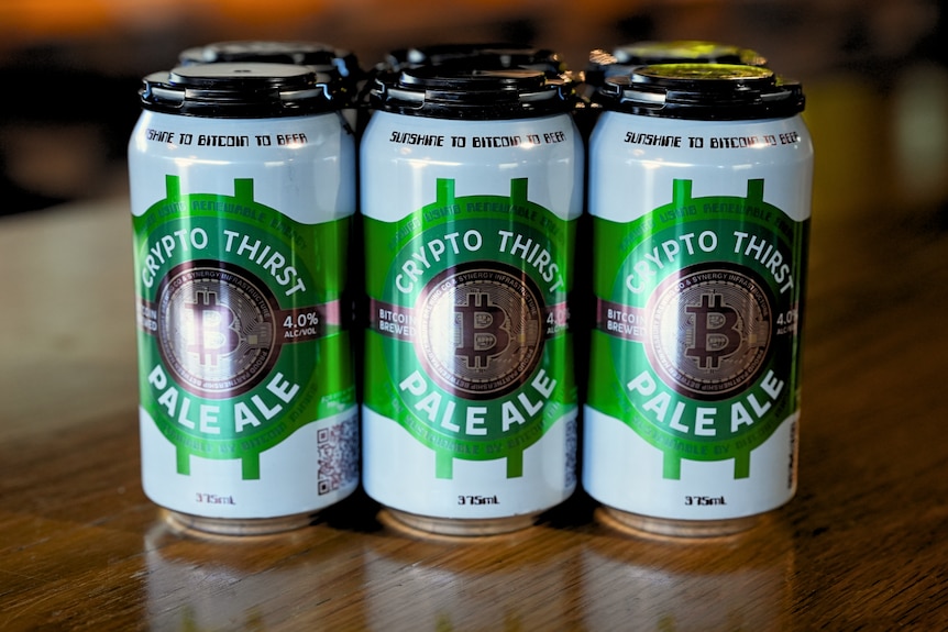 A six-pack of beer cans sitting on a wooden table. They are predominantly white with green print.
