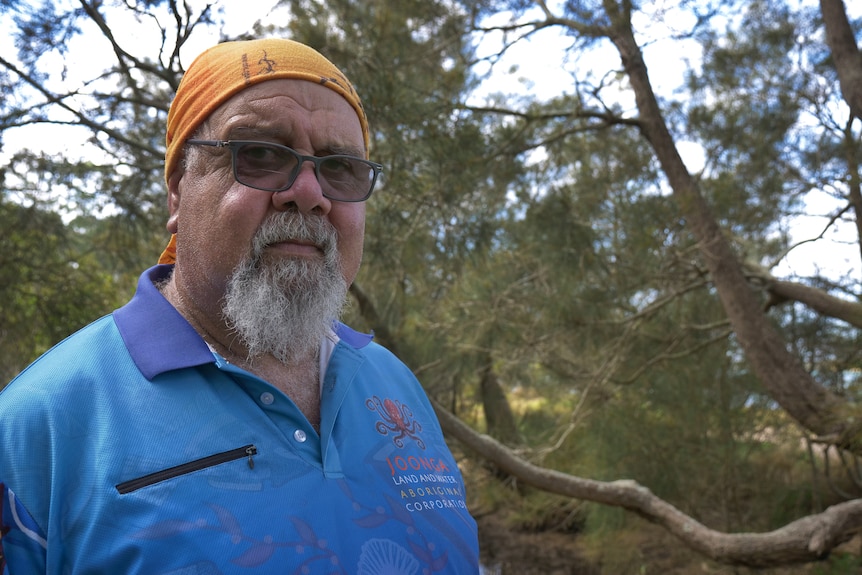 An Aboriginal man in his 60s wearing glasses stands near a tree outside and looks into the camera
