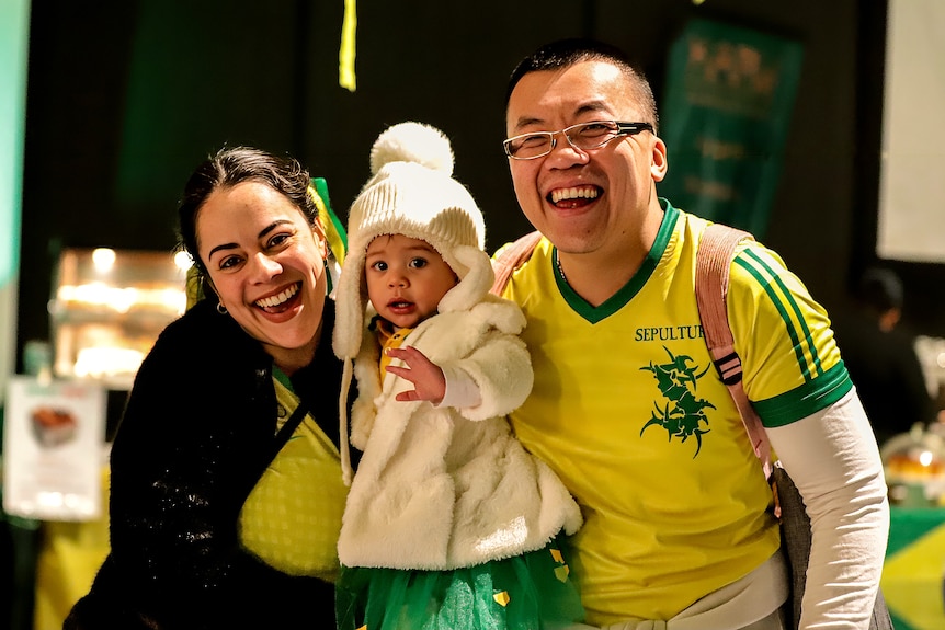 Man, woman and their young child, all wearing Brazillian yellow and green colours smile inside an event room