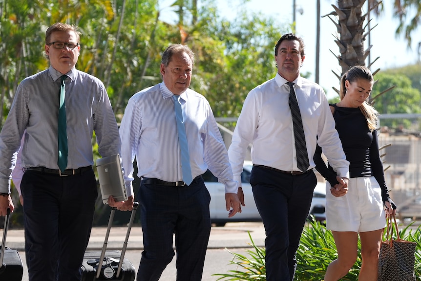 A man wearing a tie, holding his wife's hand and walking alongside two lawyers.