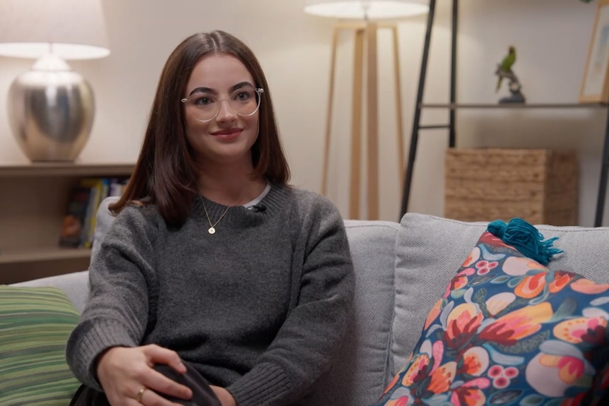 A young woman with straight, dark hair and glasses sits on a couch and smiles. She wears a grey, long-sleeved top.
