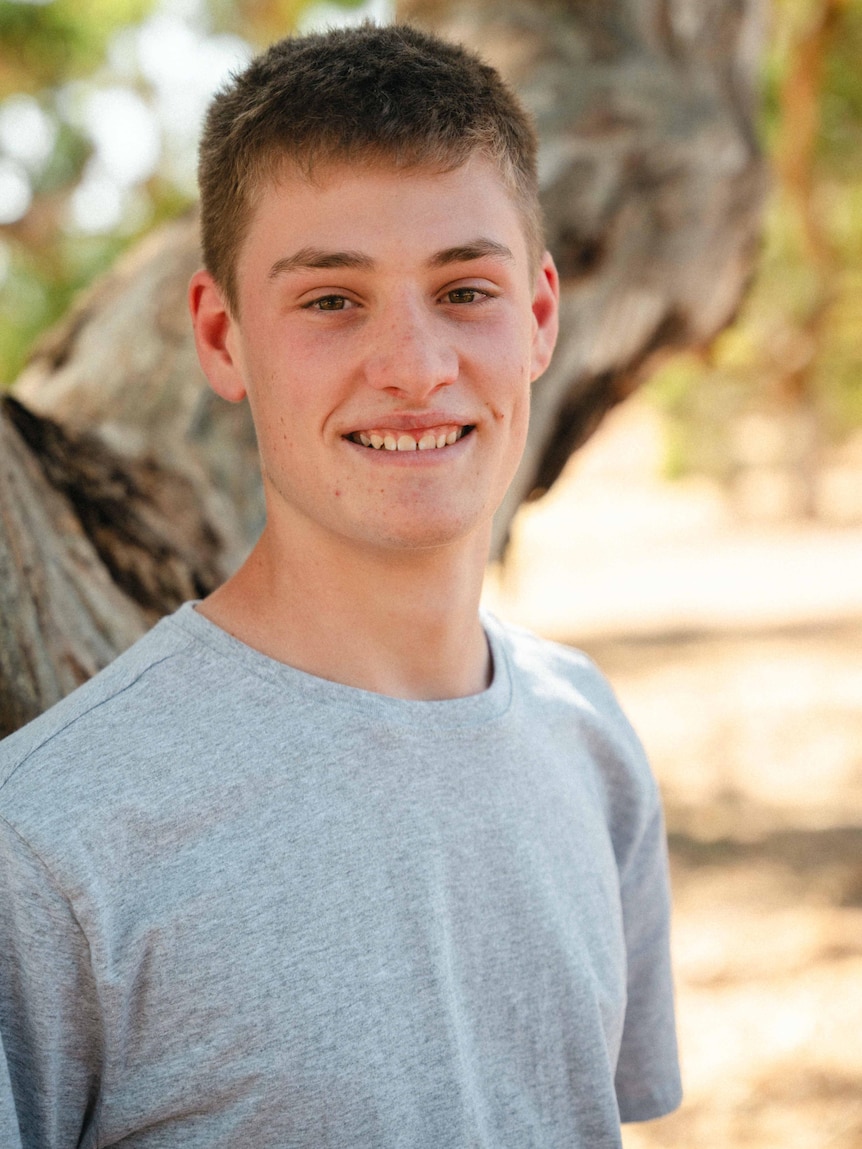 A young teen in a grey shirt standing in front of a tree with the background softened.