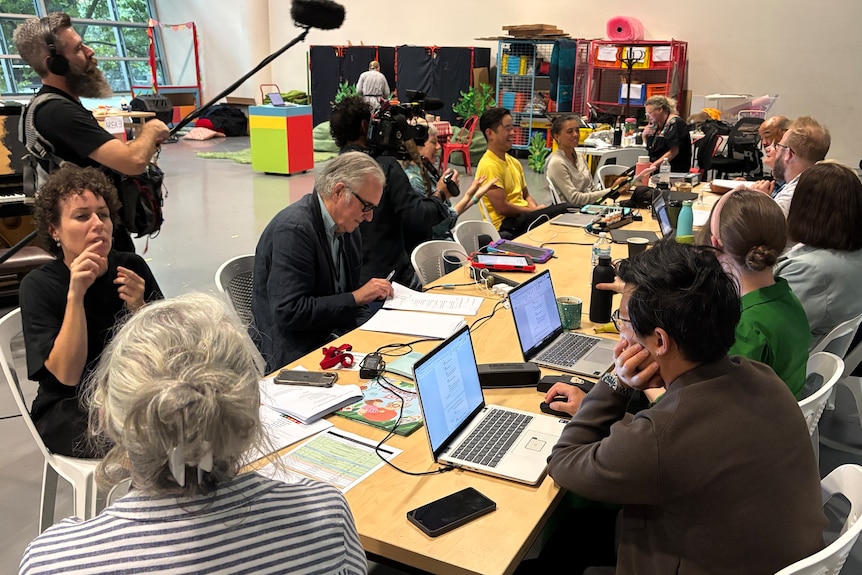 About 12 people sit around a rectangular table, some with laptops, others with paper and pen. Two camera operators are nearby