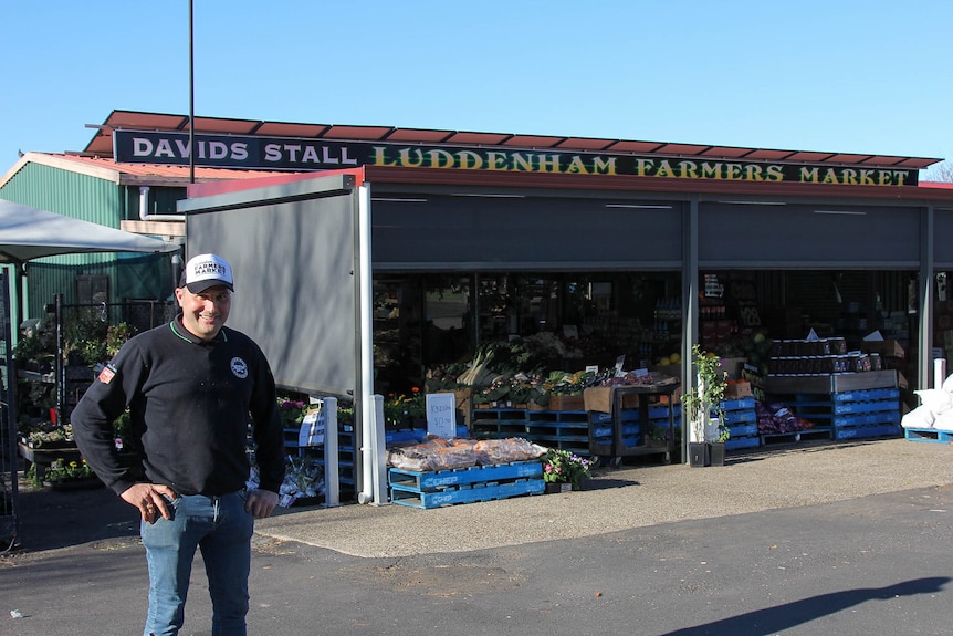 A smiling man in a cap and dark jumper stands in front of a grocery store.