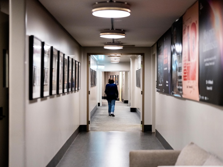 Backstage, Fran Lebowitz walks down a long hallway lined with posters and franed photos.