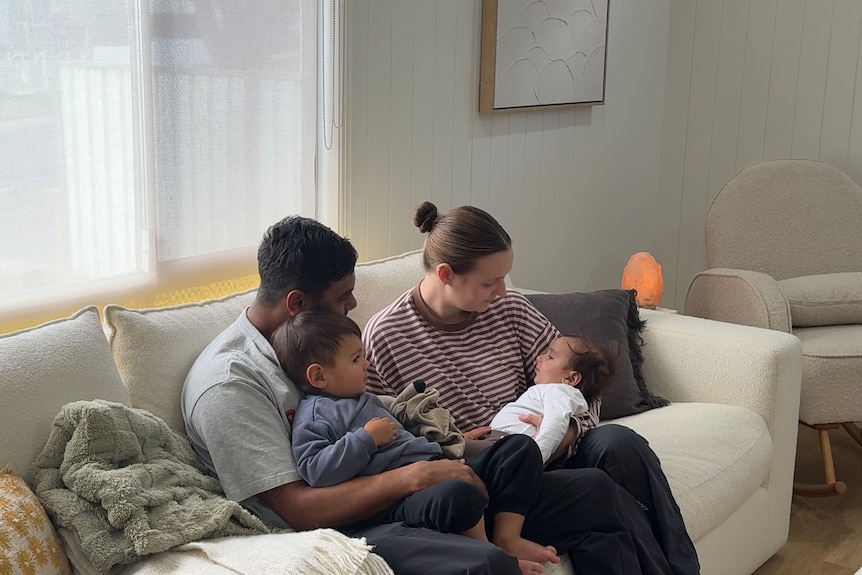 A young family sitting on couch, woman holding a baby and a man holding a toddler.