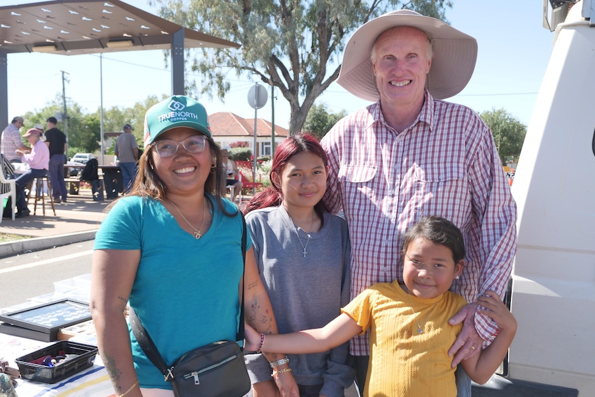A smiling woman, man and two young girls standing near an open-air market stall.