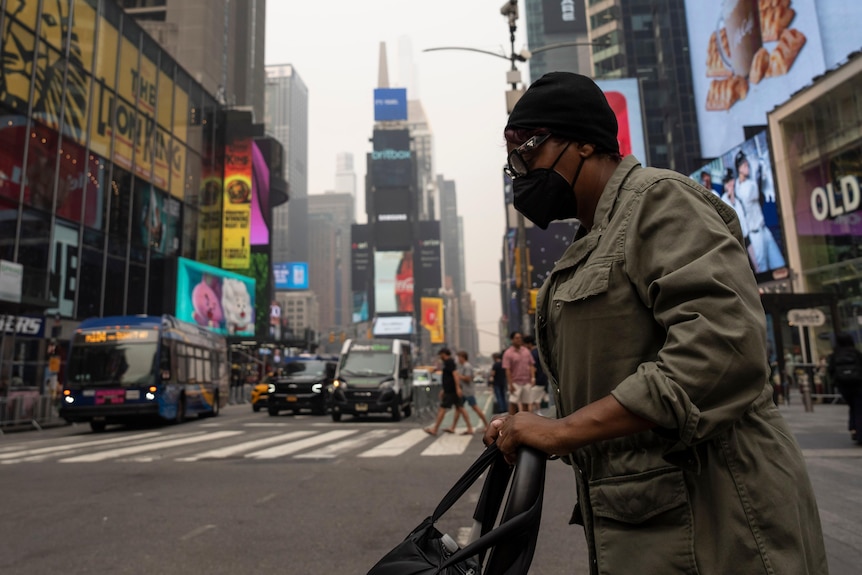 A person wearing a mask walks across a busy street in Times Square as the skyscrapers are obscured with grey haze.