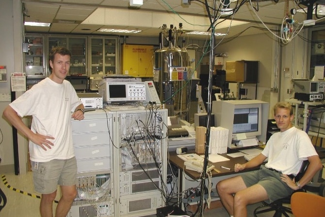 Two men in white t-shirts, one standing and one sitting either side of a machine with wires.