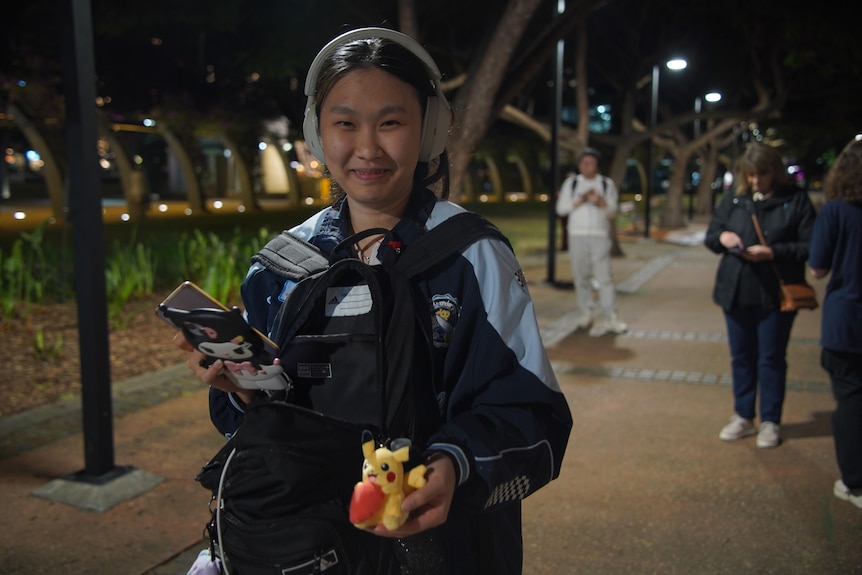 A young woman with headphones on smiles and holds a small pikachu toy in one hand