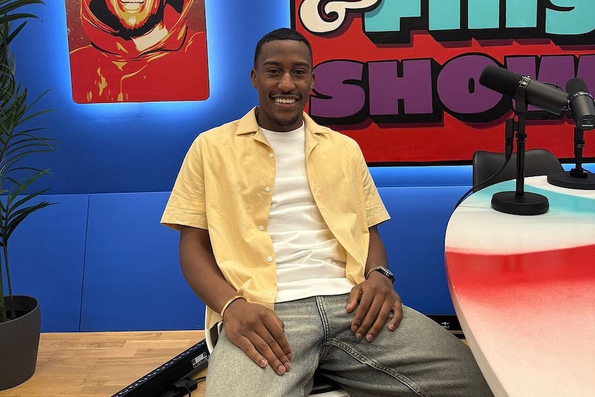 British YouTuber and rapper Yung Filly sits on a chair in a radio studio smiling, wearing yellow and white shirts and jeans.