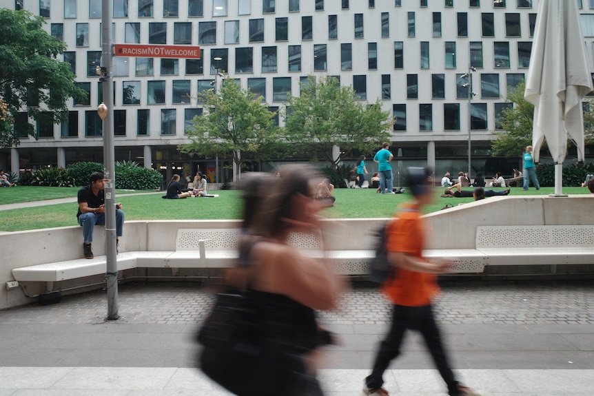 A few students walk through the UTS campus. In the background is a modern building and people lounge on a grass area.