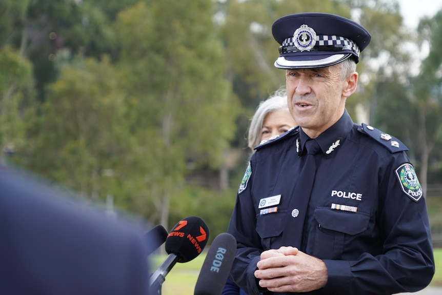 A man in police uniform stands in front of media microphones at a park
