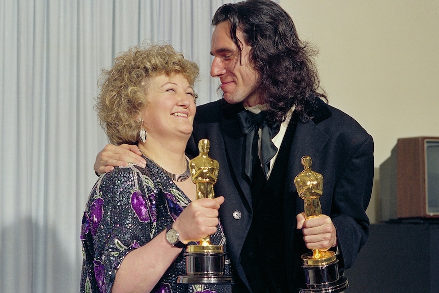 A man and a woman stand with their arms around each other holding golden trophies.