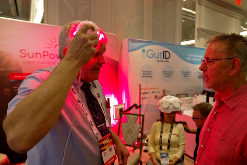 A man shining a red light to his forehead while Norman looks on, boards with information behind.