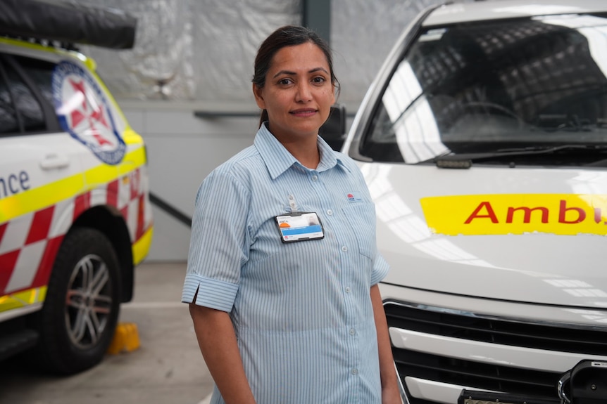 Anita Das in a uniform, smiling softly and standing in between two ambulance vehicles.