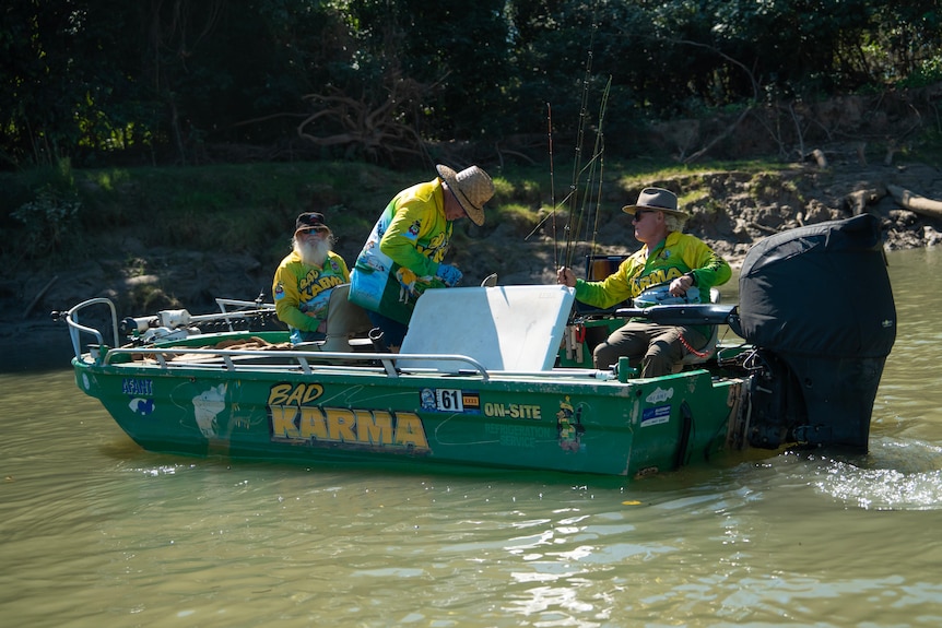 Three men on a small green fishing boat which reads 'Bad Karma'. Man on right has blonde shoulder length hair and green shirt on