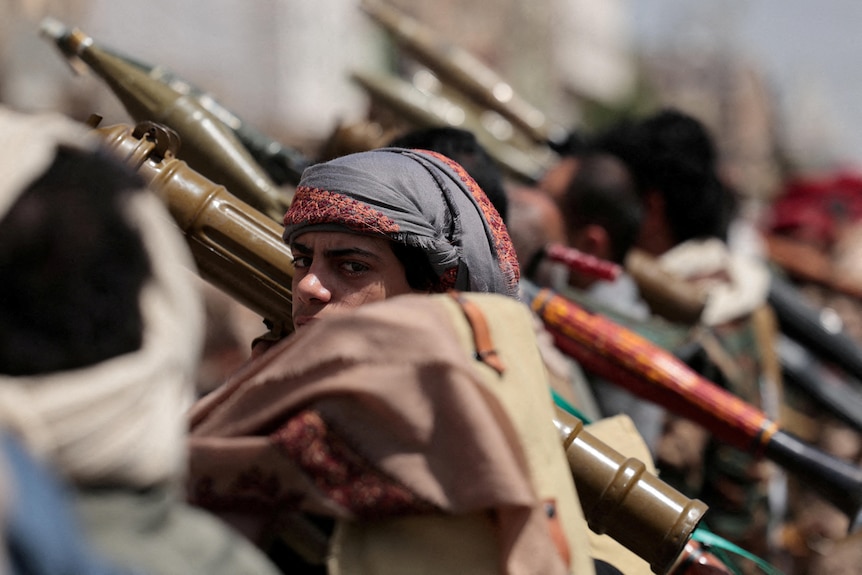 A young man with his head wrapped in cloth holding a rocket launcher over his shoulder, as he looks to the camera.