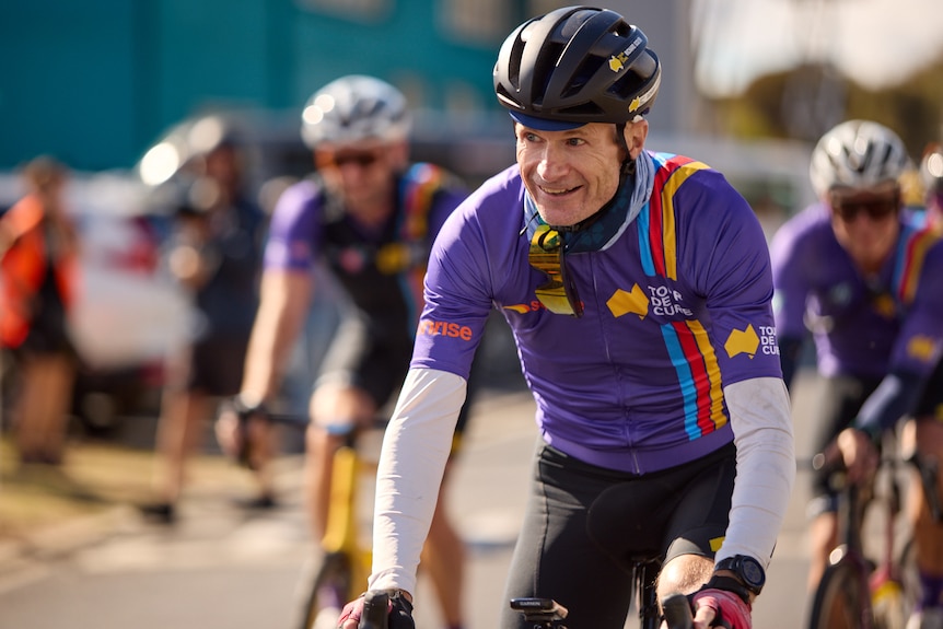 Richard Scolyer in purple and black exercise gear, standing next to his blue bike, smiling.