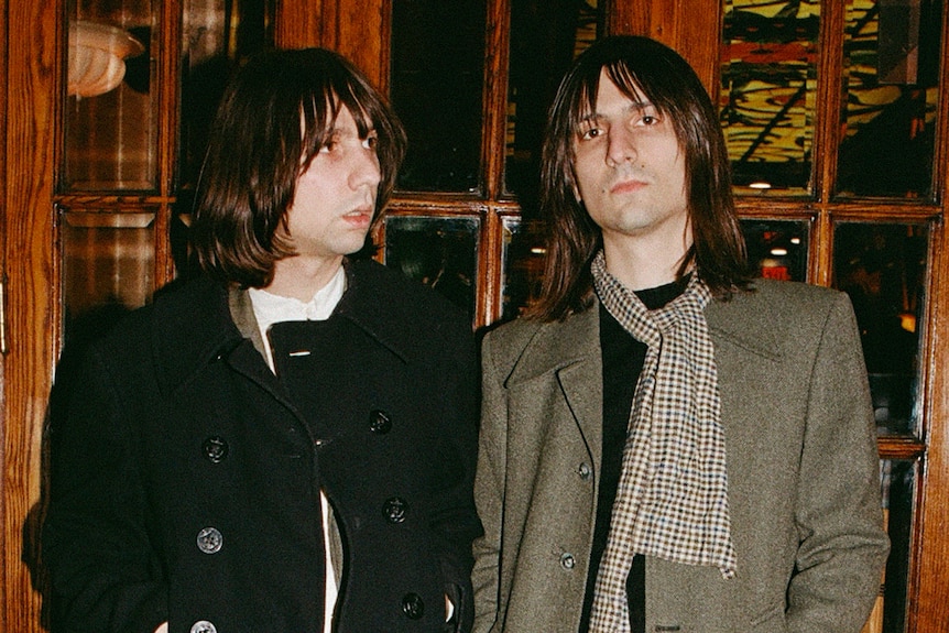 White brothers with shoulder-length brown hair pose in winter coats against a wooden window frame
