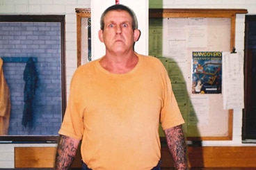 A large man in a yellow shirt and short blue shorts with tattoos on his forearms and bulging eyes